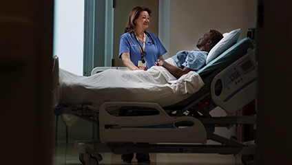 Female Vituity Medical Professional In Blue Scrubs Smiling Consulting Patient In Hospital Bed