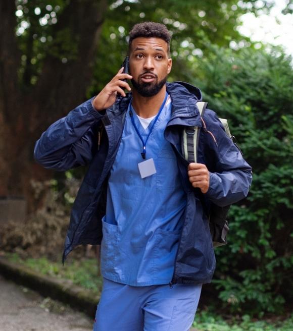Male doctor in scrubs outside walking to work with backpack talking on the phone