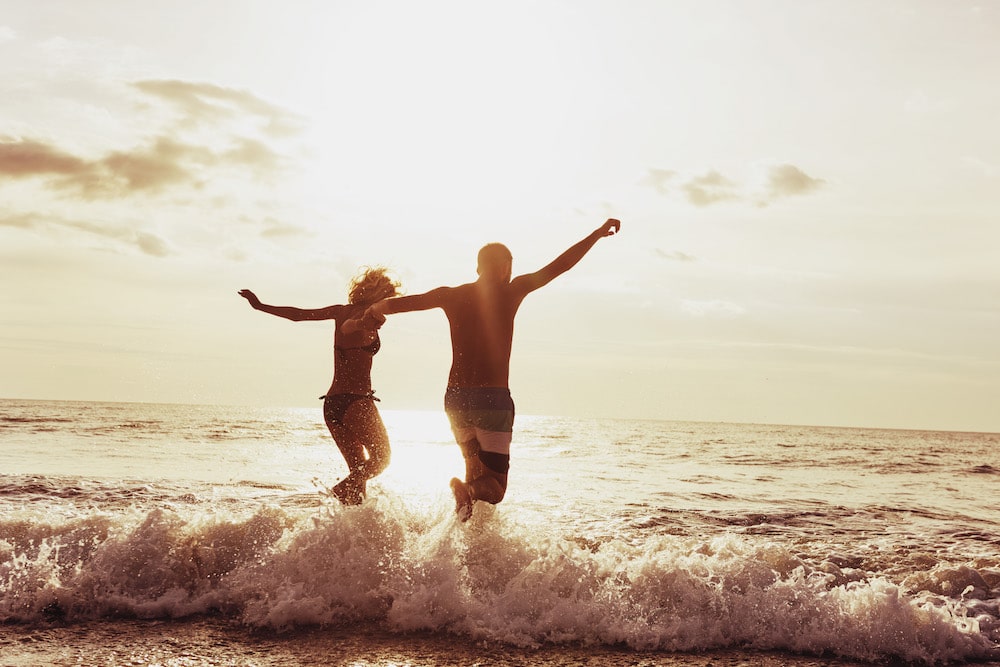 couple jumping over a wave at the beach at sunset