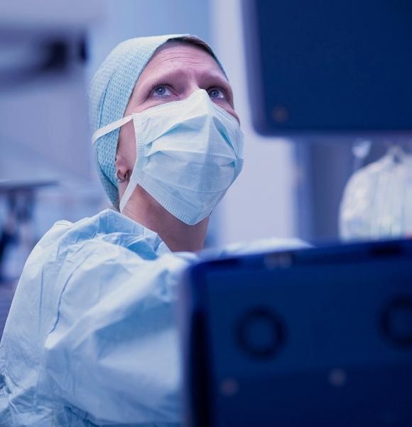 Vituity anesthesiologist wearing surgical mask and blue scrubs monitors patient vitals with urgent facial expression