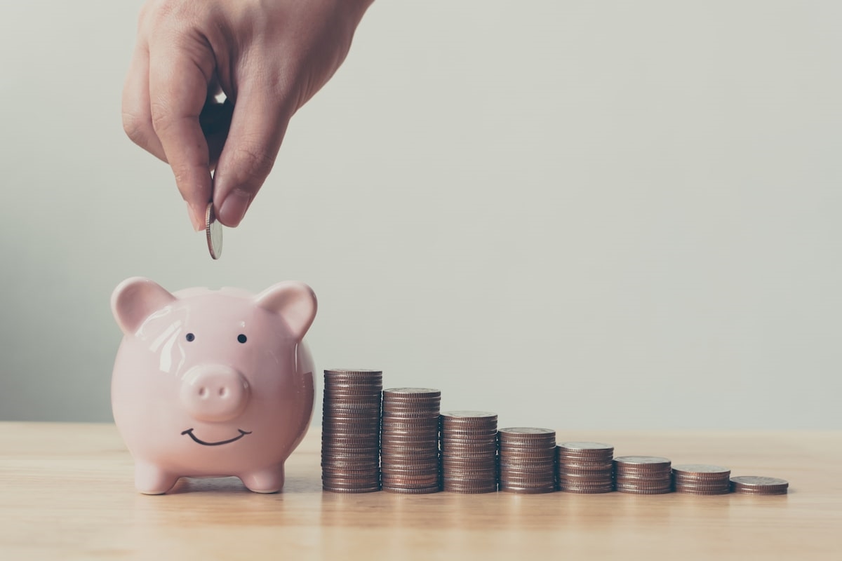 stacks of coins beside a smiling pink piggy bank with hand dropping a coin in the bank